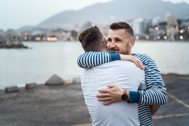 Hombre sincero abrazando pareja homosexual irreconocible mientras mira hacia otro lado contra el lago y el monte en la ciudad - foto de stock