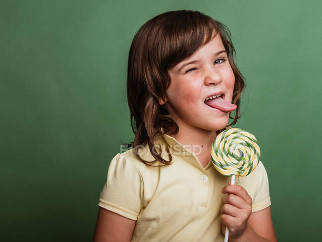 Divertido niño preadolescente lamiendo dulce remolino piruleta sobre fondo verde en el estudio y mirando a la cámara — Stock Photo