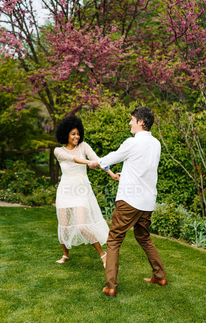 Couple multiracial optimiste tenant la main et dansant sur la pelouse tout en s'amusant dans le jardin d'été — Photo de stock