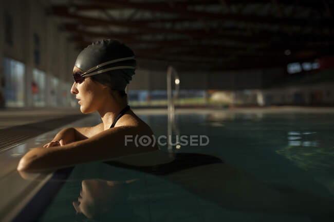 Молода красива жінка на бордюрі критого басейну, одягнена в чорний купальник, плаває у воді — стокове фото