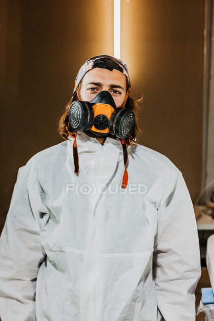 Trabalhador masculino usando respirador de segurança e traje protetor na oficina enquanto olha para a câmera — Fotografia de Stock