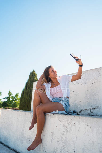 Positivo alegre joven mujer tomando uno mismo disparo en el teléfono móvil en el día soleado en verano en la ciudad - foto de stock