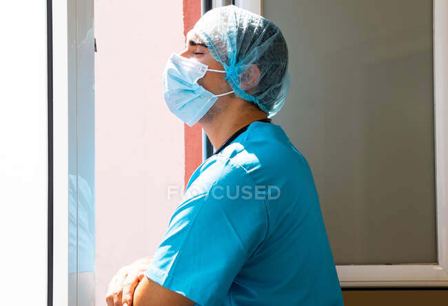 Side view of tired male medic in mask and uniform standing near window in clinic and having break — Stock Photo