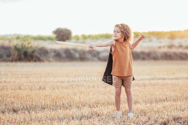 Ethnic boy in cape and with toy sword pretending being knight and playing in field in summer — Stock Photo