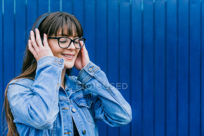 Encantada mulher ouvindo música em fones de ouvido e desfrutando de músicas com olhos fechados em fundo azul na cidade — Fotografia de Stock