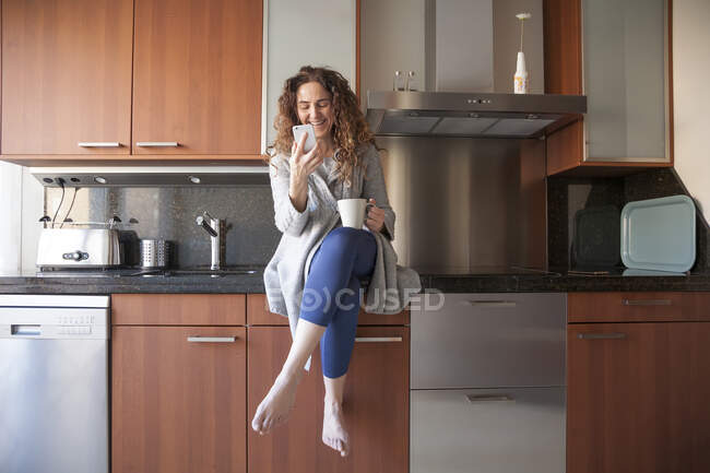 Деловая женщина с вьющимися волосами сидит на кухне, принимая вливание, используя смартфон и работая дома — стоковое фото