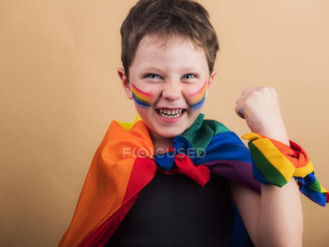 Cheerful kid with makeup on cheeks with LGBTQ flag while looking at camera on beige background — Stock Photo