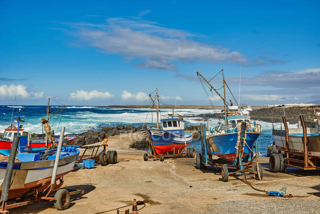 Buques coloridos ubicados en remolques corroídos en la costa cerca del mar ondeando contra el cielo azul nublado en Fuerteventura, España - foto de stock