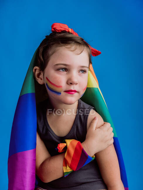 Kleines Mädchen mit gebundener Regenbogenfahne auf dem Kopf schaut vor blauem Hintergrund weg — Stockfoto