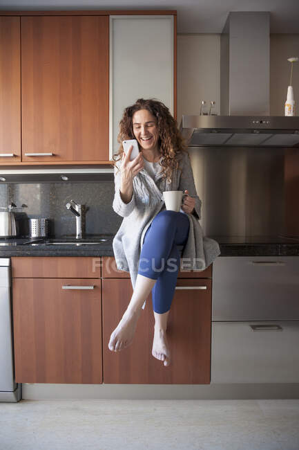 Femme d'affaires avec les cheveux bouclés assis dans la cuisine prendre une infusion tout en utilisant son smartphone et de travailler à la maison — Photo de stock