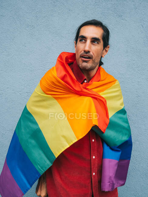 Homosexuelle ethnische Männer in Schutzmaske und in Regenbogenflagge gehüllt schauen in die Kamera gegen eine graue Wand in der Stadt — Stockfoto