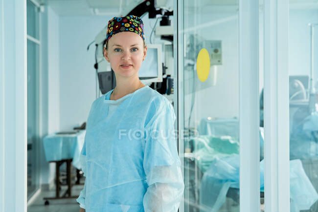 Médico adulto seguro de sí mismo en gorra médica ornamental mirando a la cámara contra la pared de vidrio en el hospital - foto de stock