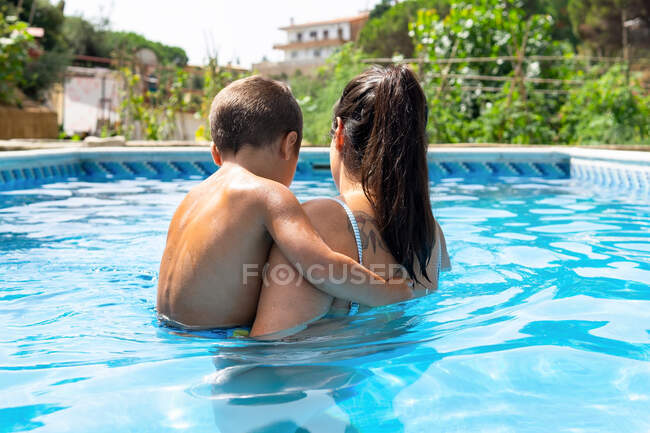 Обратный вид на неузнаваемую мать с сыном, стоящим в бассейне возле зеленых деревьев в солнечный летний день — стоковое фото
