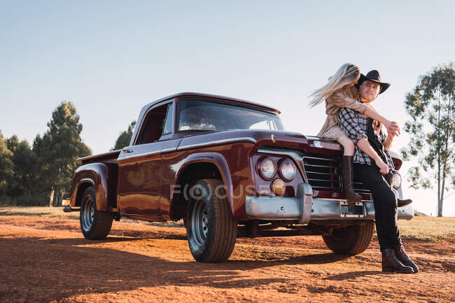 Angolo basso di coppia alla moda in amore abbracciare vicino parcheggiato rosso retro pick-up auto nella giornata di sole in estate — Foto stock
