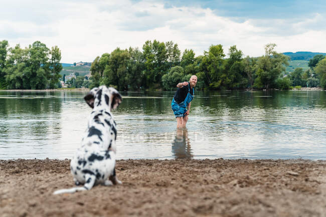 Hombre feliz con barba en ropa casual jugando con el cachorro mientras está de pie en el agua del lago en verano - foto de stock