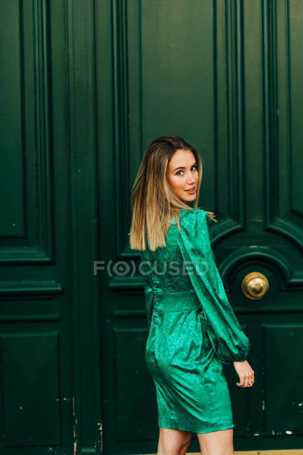 Mulher alegre em vestido verde elegante em pé perto de portas de madeira ornamentais na rua e olhando para a câmera — Fotografia de Stock