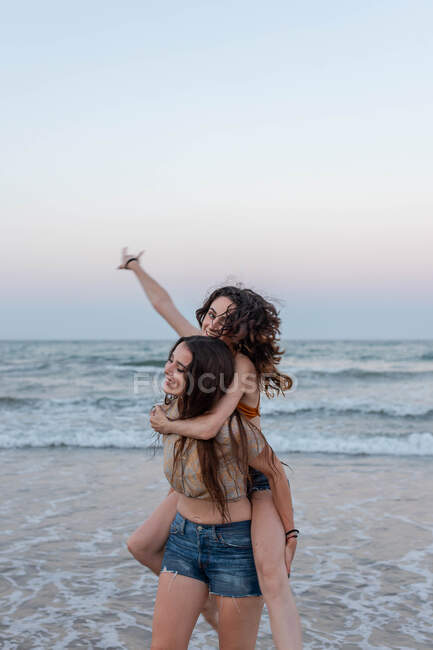 Young woman giving piggyback ride to girlfriend while standing in waving sea water in evening — Stock Photo