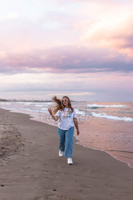 Mulher jovem positivo em óculos de sol na moda e roupa elegante andando à beira-mar contra o mar na noite de verão — Fotografia de Stock