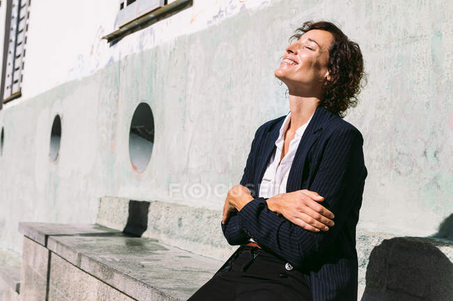 Trabajadora positiva con ropa elegante abrazándose sentada en un banco de hormigón y disfrutando de rayos de luz solar con los ojos cerrados - foto de stock