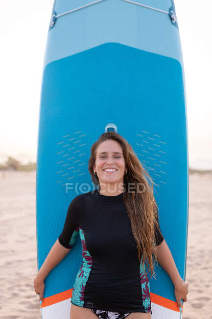 Glückliche Surferin steht im Sommer mit blauem SUP-Board am Sandstrand und blickt in die Kamera — Stockfoto