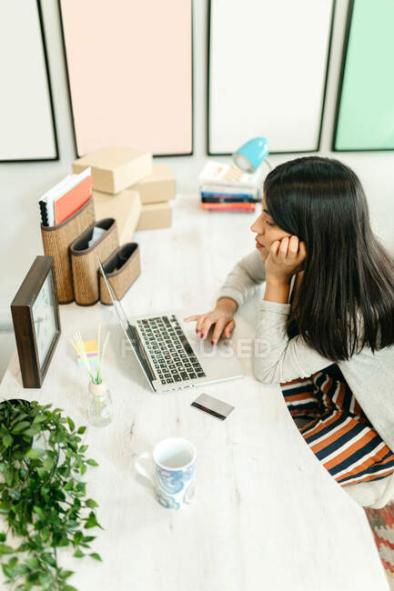 Vista lateral desde arriba de la hembra usando netbook durante las compras en línea mientras está sentado en la mesa con tarjeta de plástico en casa y la elección de productos - foto de stock