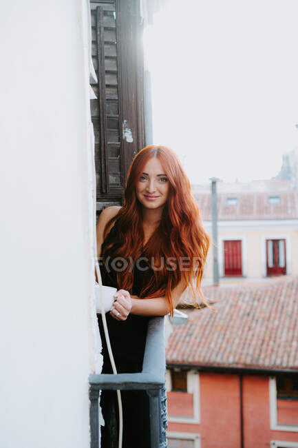 Zufriedene Frau mit roten Haaren steht mit Getränkebecher auf Balkon und blickt in die Kamera — Stockfoto