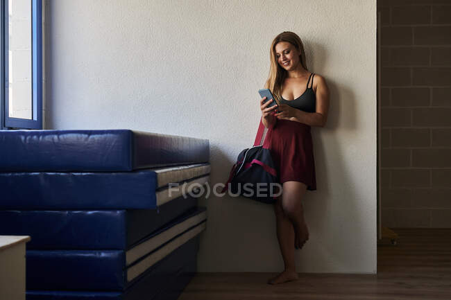 Atleta feminina alegre em pé perto do tapete com saco esportivo no ginásio e no telefone móvel de navegação — Fotografia de Stock