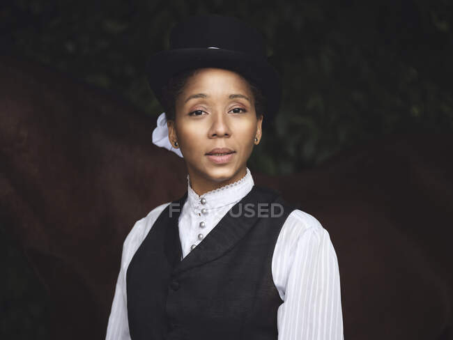 Grave donna afroamericana adulta in abito elegante e cappello guardando la fotocamera vicino a cavallo marrone e piante verdi alla luce del giorno — Foto stock