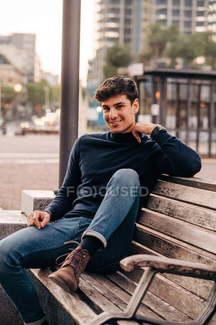 Delighted male wearing stylish clothes sitting on wooden bench in street and looking at camera — Stock Photo