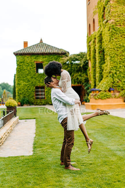 Side view of man lifting black woman while standing on meadow in garden against building with ivy on wall — Stock Photo