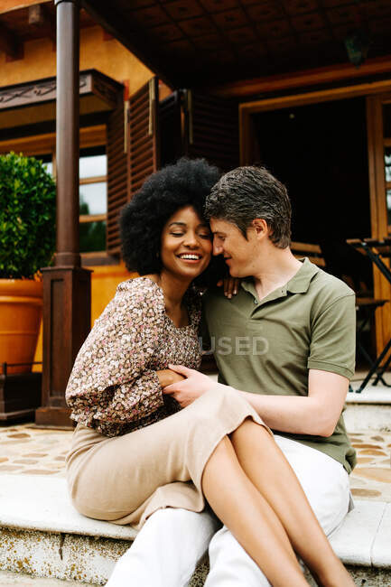 Positive multiracial couple sitting on stone stairs of porch of cottage and embracing while enjoying weekend together — Stock Photo
