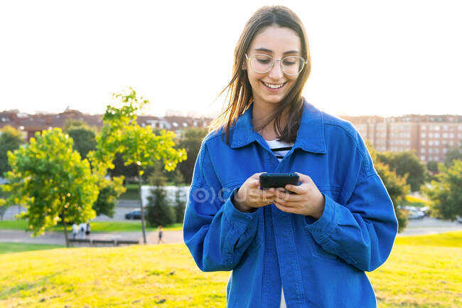 Стильна жінка стоїть на зеленому трав'яному пагорбі і переглядає на смартфоні в сонячний день — стокове фото