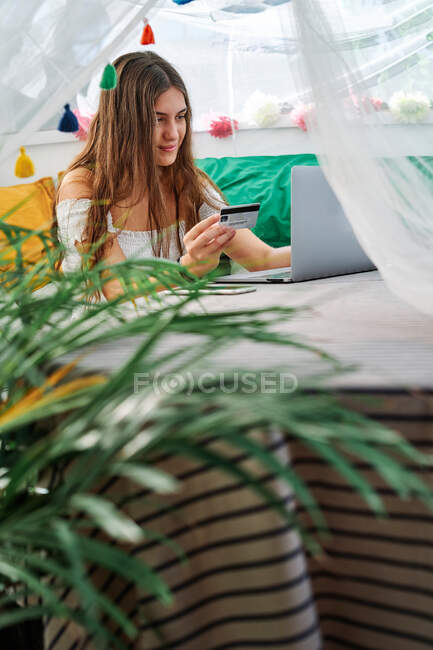 Задоволена жінка сидить за столом з ноутбуком і робить покупки з пластиковою карткою під час онлайн-покупки в наметі на подвір'ї — стокове фото