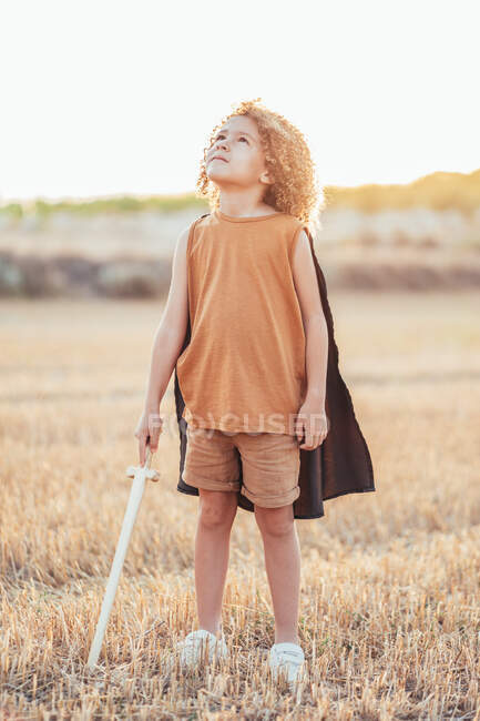 Ethnic boy in cape and with toy sword pretending being knight and playing in field in summer — Stock Photo