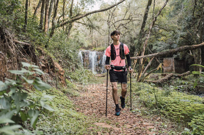 Male traveler with trekking poles walking along trail near waterfall in tropical forest — Stock Photo