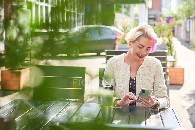 Donna alternativa con capelli corti che naviga sui social media sullo smartphone mentre siede a tavola nel caffè di strada nella giornata di sole — Foto stock