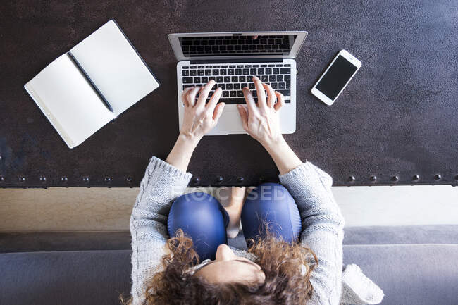 Вид сверху на деловую женщину с кудрявыми волосами, сидящую на диване и работающую с ноутбуком — стоковое фото