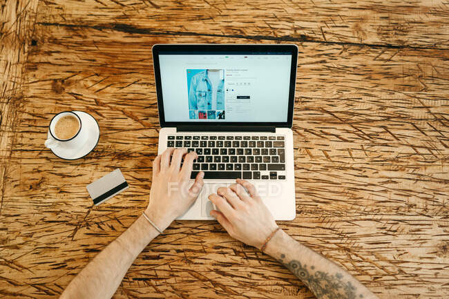 Вид зверху на урожай анонімний чоловік, який вибирає одяг на сайті під час онлайн-покупки, використовуючи нетбук і сидячи за столом з пластиковою карткою в кафе — стокове фото