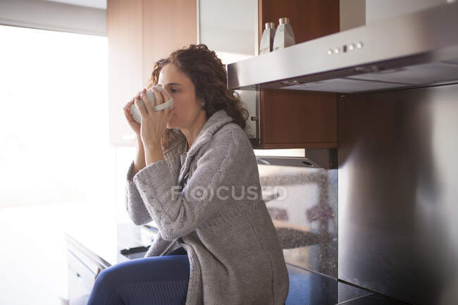 Vue latérale de la femme aux cheveux bouclés assise dans la cuisine prenant une infusion — Photo de stock