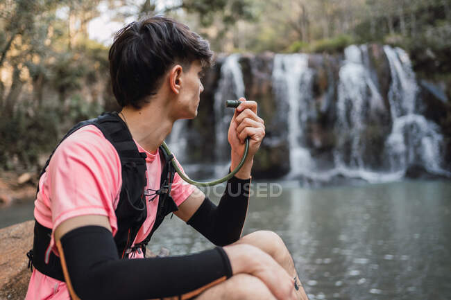 Seitenansicht des männlichen Wanderers mit Hydratationsrucksack, der aus dem Schlauch trinkt, während er beim Trekking im Wald Pause macht und den Blick auf den See in der Nähe des Wasserfalls bewundert — Stockfoto