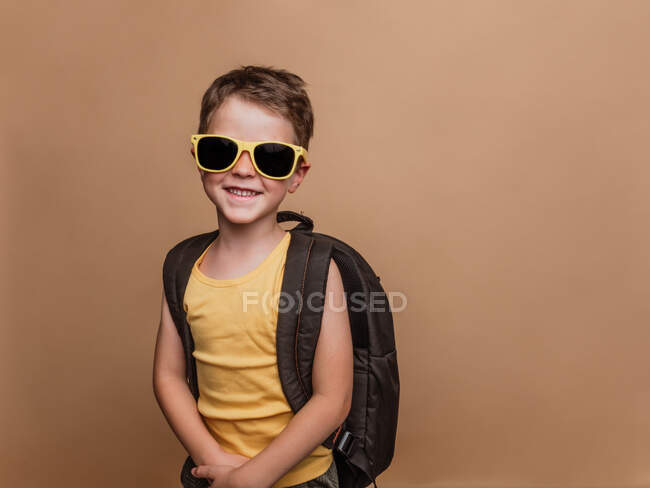 Positivo legal pré-adolescente estudante em óculos de sol e com mochila olhando para a câmera no fundo marrom em estúdio — Fotografia de Stock