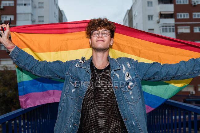 From below of happy homosexual male standing with rainbow flag in raised arms on bridge and closed eyes — Stock Photo