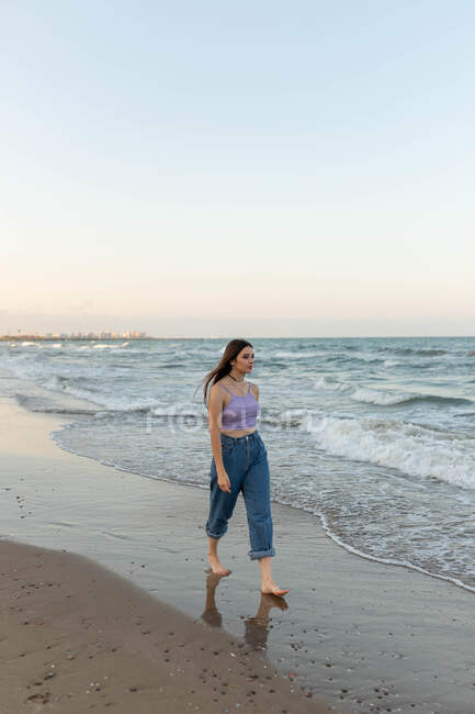Joven mujer descalza en la parte superior y pantalones vaqueros caminando sobre arena mojada cerca del mar ondeando por la noche en la playa - foto de stock