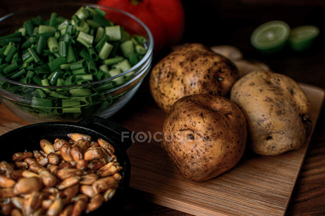 High angle of raw potatoes and chopped spring onion placed on wooden cutting board near bowl with grains prepared for cooking chicken broth — Stock Photo