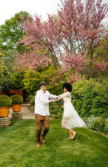 Оптимістична багаторасова пара тримає руки і танцює на газоні, розважаючись в літньому саду — стокове фото
