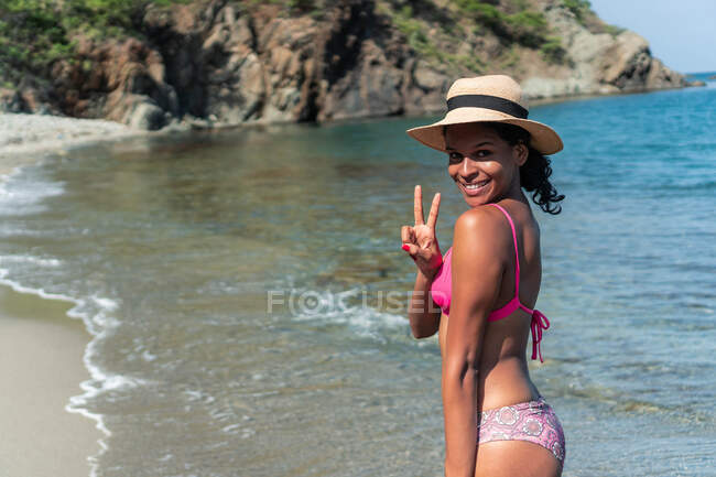 Вид збоку весела етнічна жінка-туристка в купальнику і капелюсі, дивлячись на камеру на узбережжі океану, поки вона робить символ перемоги — стокове фото