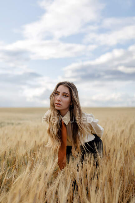 Giovane femmina con i capelli ondulati guardando la fotocamera piegarsi in avanti nel campo di campagna sotto cielo nuvoloso su sfondo sfocato — Foto stock