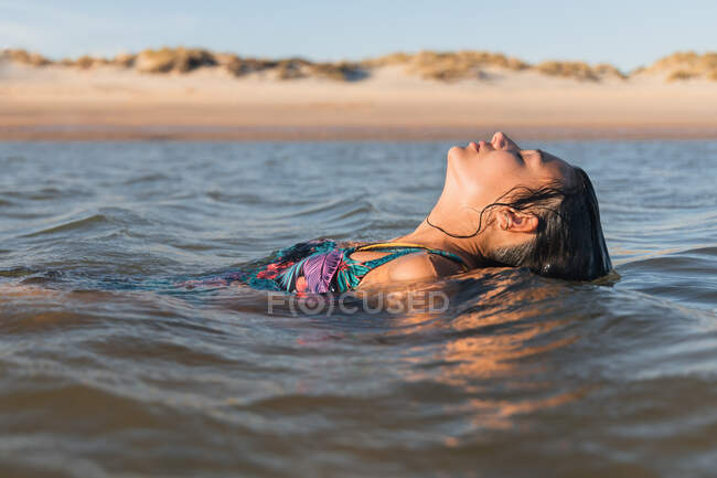 Mujer serena con el pelo mojado nadando en el mar tranquilo en la noche de verano y los ojos cerrados - foto de stock