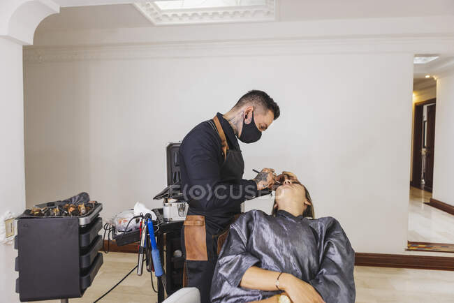 Homem em máscara manchando fundação de cara de mulher loira durante o trabalho em estúdio de maquiagem profissional — Fotografia de Stock