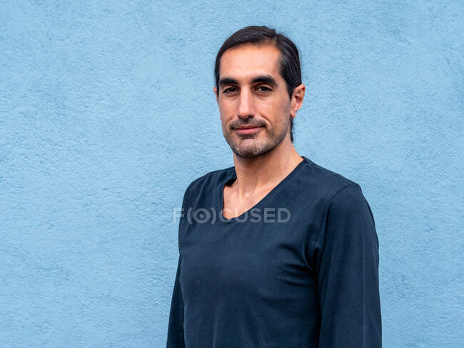 Handsome ethnic male standing against light blue wall in city street and looking at camera — Stock Photo